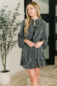 Woman wearing a black and white patterned dress standing indoors with a plant in the background